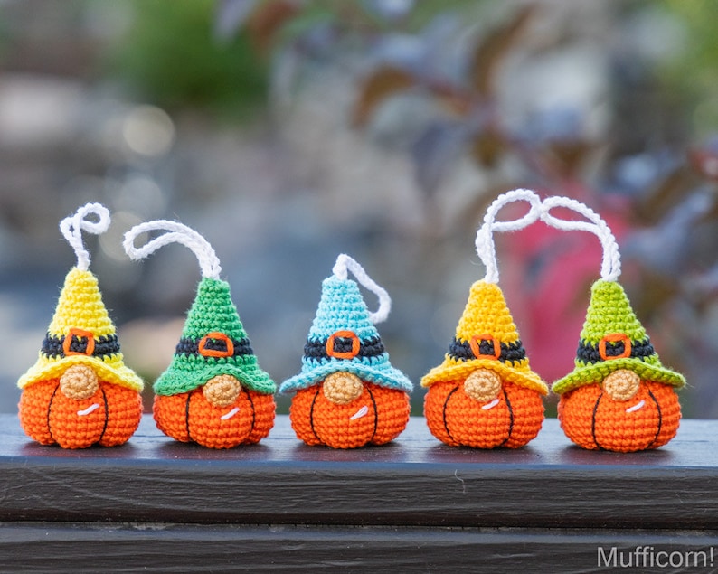 May include: Five crocheted pumpkin gnomes with different colored hats. The gnomes are wearing black belts and have orange pumpkin bodies. The gnomes are hanging from white strings.