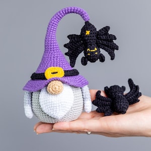 Puede incluir: Un gnomo de crochet morado que lleva un sombrero negro con una hebilla amarilla. El gnomo tiene una barba blanca y sostiene un murci&eacute;lago de crochet negro. Hay otro murci&eacute;lago de crochet negro a la derecha del gnomo.