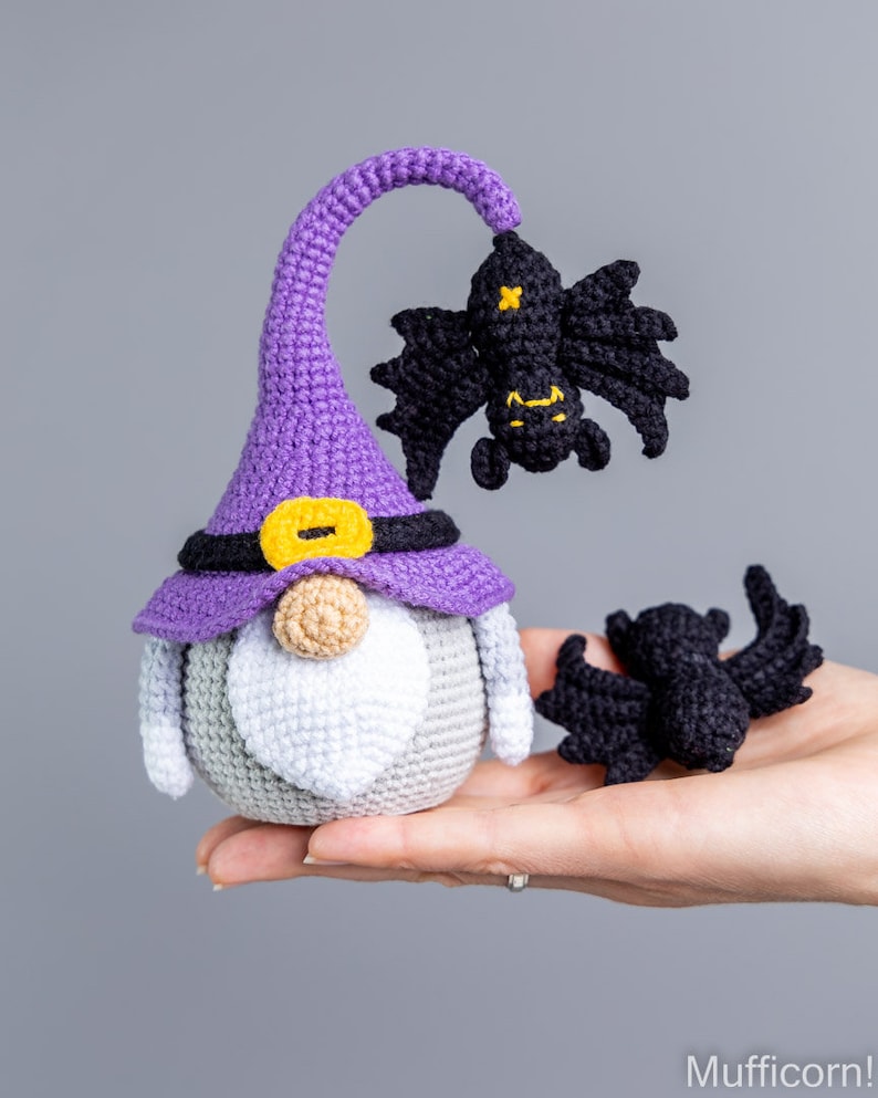 Puede incluir: Un gnomo de crochet morado que lleva un sombrero negro con una hebilla amarilla. El gnomo tiene una barba blanca y sostiene un murci&eacute;lago de crochet negro. Hay otro murci&eacute;lago de crochet negro a la derecha del gnomo.