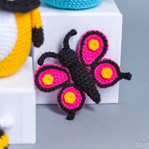 May include: A pink and black crocheted butterfly with yellow accents. The butterfly is sitting on a white surface.