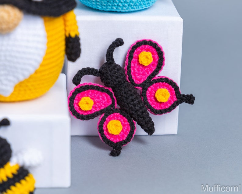 May include: A pink and black crocheted butterfly with yellow accents. The butterfly is sitting on a white surface.