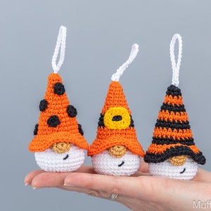 May include: Three handmade crochet gnome ornaments. Each gnome has a white body, a beige beard, and a pointed orange hat. One hat has black polka dots, another has a yellow buckle, and the third has black stripes. Each has a white loop for hanging.