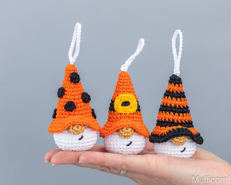 May include: Three handmade crochet gnome ornaments. Each gnome has a white body, a beige beard, and a pointed orange hat. One hat has black polka dots, another has a yellow buckle, and the third has black stripes. Each has a white loop for hanging.