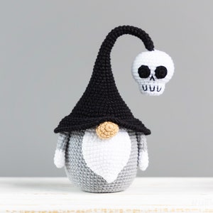 May include: A gray and white crocheted gnome wearing a black witch hat with a white skull attached to the top. The skull has black eyes and a toothy grin.
