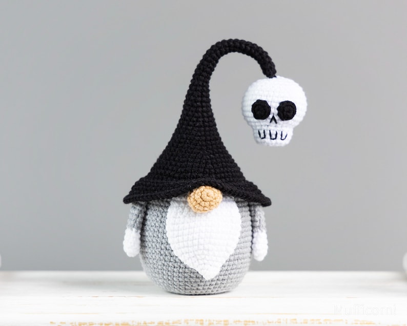 May include: A gray and white crocheted gnome wearing a black witch hat with a white skull attached to the top. The skull has black eyes and a toothy grin.