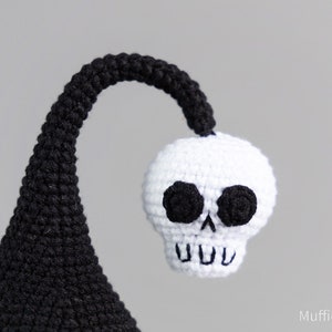 May include: A black and white crocheted skull with black eyes and a toothy grin. The skull is attached to a black crocheted stem.
