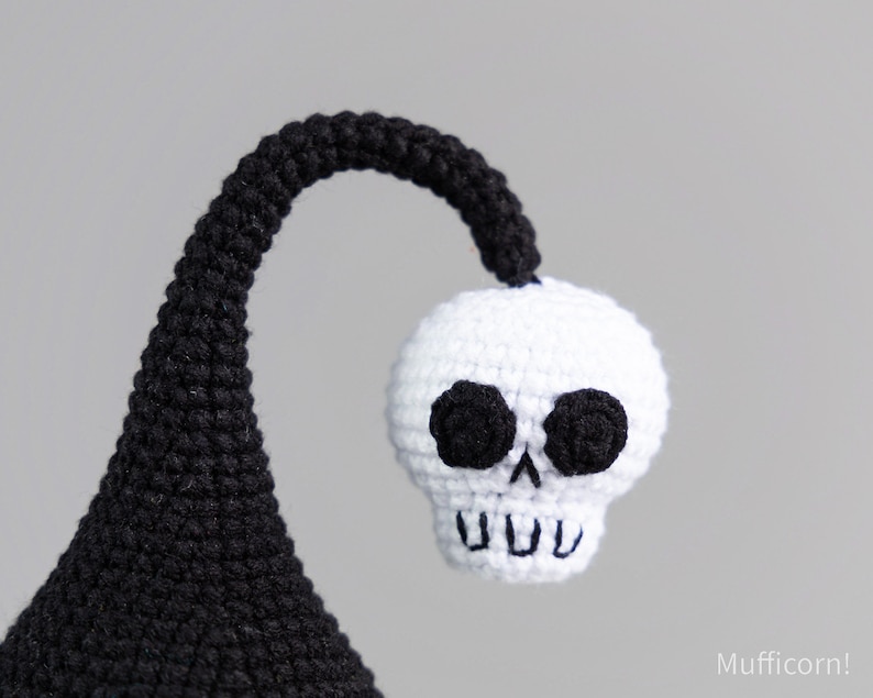May include: A black and white crocheted skull with black eyes and a toothy grin. The skull is attached to a black crocheted stem.