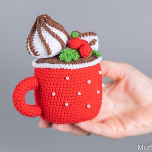 May include: A crocheted red mug with a white rim and white pearl accents. The mug is filled with a brown crocheted chocolate drink topped with white crocheted whipped cream, a brown crocheted chocolate drizzle, and red crocheted berries.