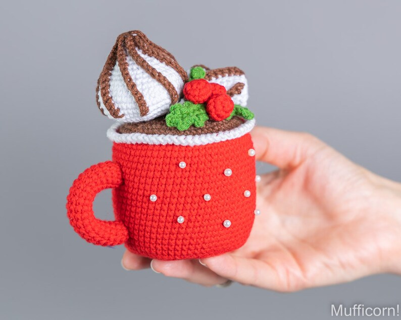 May include: A crocheted red mug with a white rim and white pearl accents. The mug is filled with a brown crocheted chocolate drink topped with white crocheted whipped cream, a brown crocheted chocolate drizzle, and red crocheted berries.
