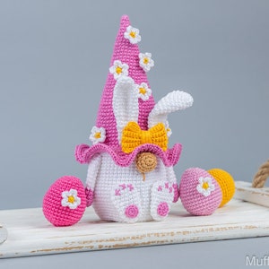 May include: A pink crocheted Easter gnome with a white bunny face, yellow bow, and white flowers on its hat. The gnome is sitting on a white wooden tray with two pink crocheted Easter eggs.