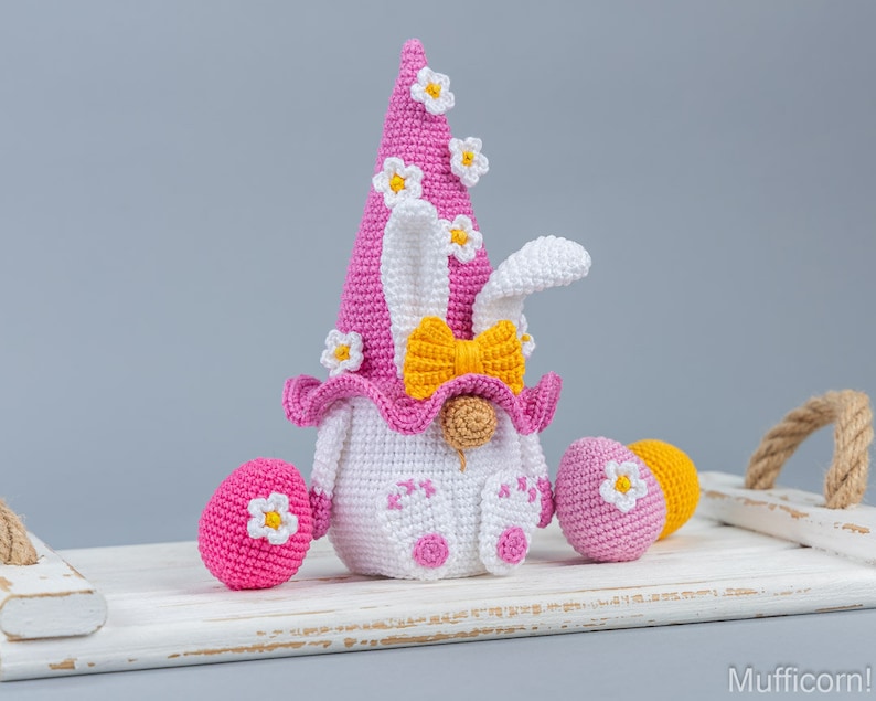May include: A pink crocheted Easter gnome with a white bunny face, yellow bow, and white flowers on its hat. The gnome is sitting on a white wooden tray with two pink crocheted Easter eggs.