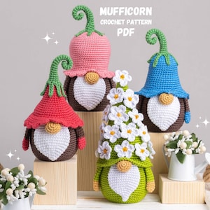 Könnte beinhalten: Vier gehäkelte Gnome, jeder mit einem Hut in einer anderen Farbe. Die Gnome stehen auf Holzblöcken. Die Gnome tragen weiße Bärte und haben braune Körper. Die Gnome sind von weißen Blumen und einer kleinen weißen Gießkanne umgeben.