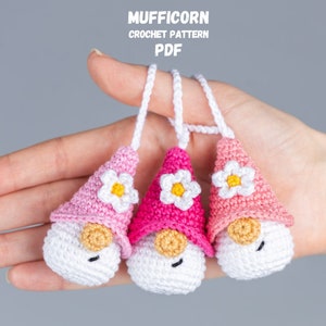 May include: Three handmade crochet gnomes in shades of pink, white, and tan. Each gnome has a conical hat with a white flower, a white body, and a tan nose. The gnomes are suspended by white cords. Text at the top reads "MUFFICORN CROCHET PATTERN PDF".