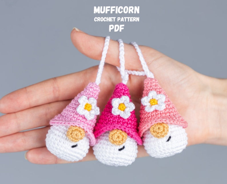 May include: Three handmade crochet gnomes in shades of pink, white, and tan. Each gnome has a conical hat with a white flower, a white body, and a tan nose. The gnomes are suspended by white cords. Text at the top reads "MUFFICORN CROCHET PATTERN PDF".