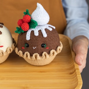May include: Two crocheted Christmas pudding ornaments on a wooden tray. The pudding on the left is white with chocolate drizzle and the pudding on the right is brown with white icing and a red holly sprig.