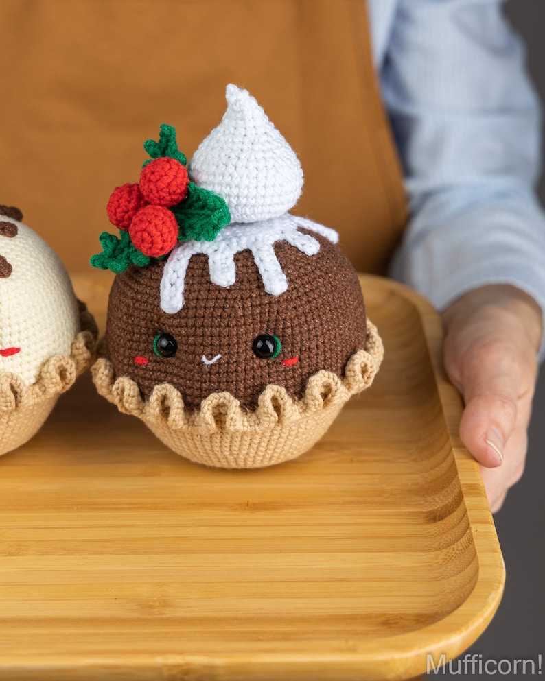 May include: Two crocheted Christmas pudding ornaments on a wooden tray. The pudding on the left is white with chocolate drizzle and the pudding on the right is brown with white icing and a red holly sprig.