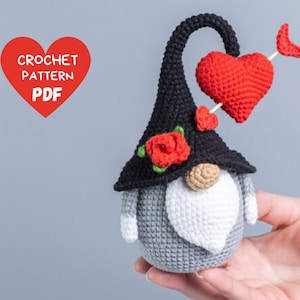 May include: A crocheted gnome wearing a black hat with a red rose and a heart-shaped arrow. The gnome has a white beard and a grey body.