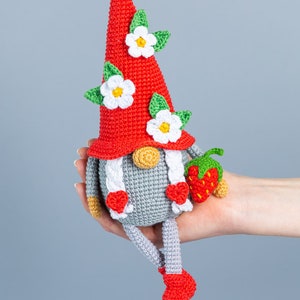 Op de afbeelding: Een gehaakte gnome met een rode muts, witte bloemen en groene bladeren. De gnome houdt een rode aardbei vast en heeft grijze benen en rode schoenen.