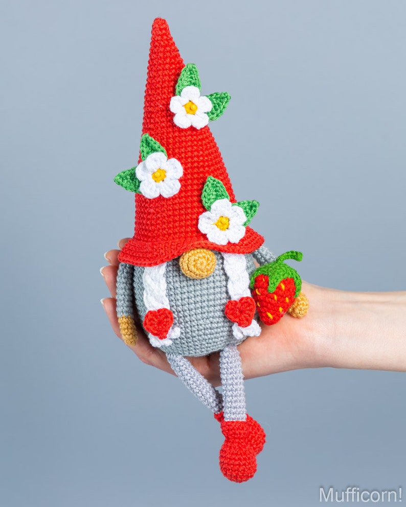 Op de afbeelding: Een gehaakte gnome met een rode muts, witte bloemen en groene bladeren. De gnome houdt een rode aardbei vast en heeft grijze benen en rode schoenen.