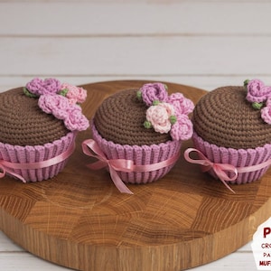 Puede incluir: Tres juguetes de cupcake de crochet con glaseado rosa y decoraciones de flores rosas. Los cupcakes están sobre una superficie de madera.