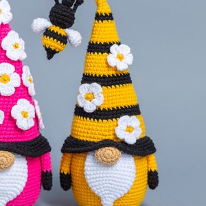 May include: Two crocheted gnomes, one yellow and one pink, with white flower accents. The yellow gnome has a black and yellow striped hat with a crocheted bee on top.