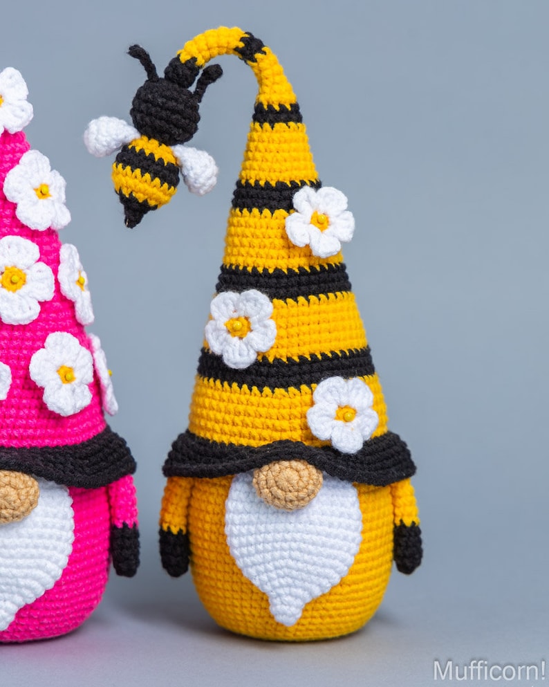 May include: Two crocheted gnomes, one yellow and one pink, with white flower accents. The yellow gnome has a black and yellow striped hat with a crocheted bee on top.