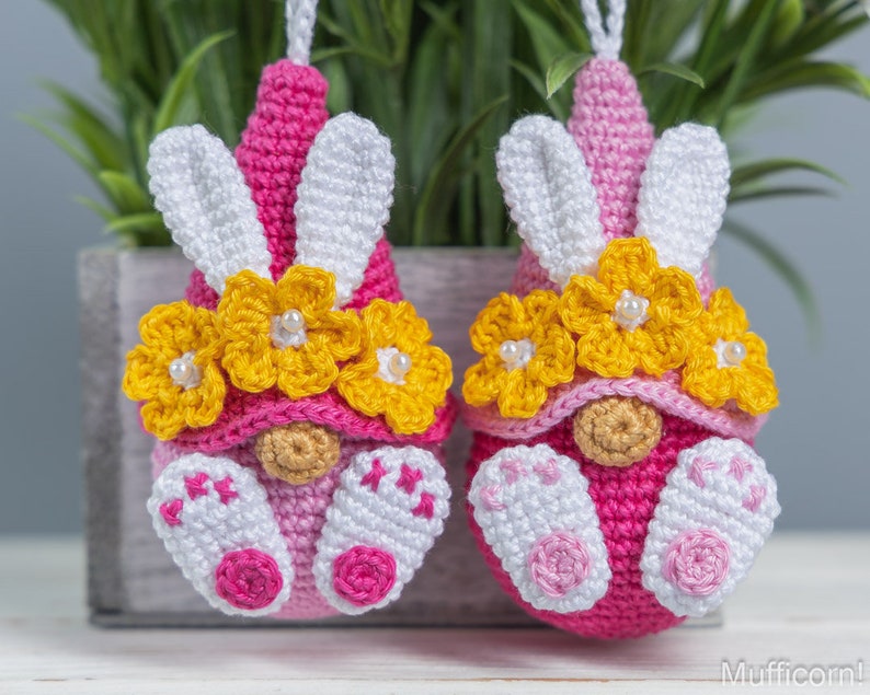 May include: Two pink crocheted gnomes with white bunny ears and yellow flower crowns. The gnomes are wearing pink and white crocheted outfits.