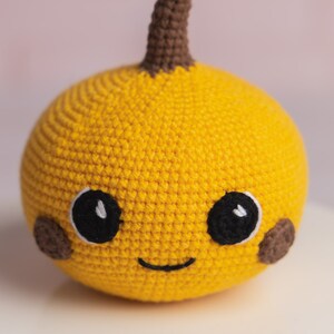 May include: A crocheted yellow pumpkin with a brown stem and a smiling face. The pumpkin has black eyes and brown cheeks.