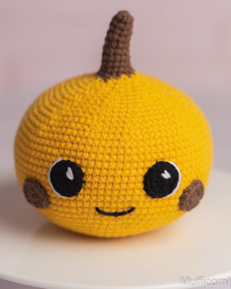 May include: A crocheted yellow pumpkin with a brown stem and a smiling face. The pumpkin has black eyes and brown cheeks.