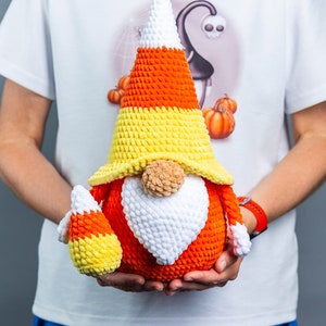 May include: A crocheted candy corn gnome with a white beard and a yellow, orange, and white candy corn hat. The gnome is holding a smaller candy corn gnome in its hands.