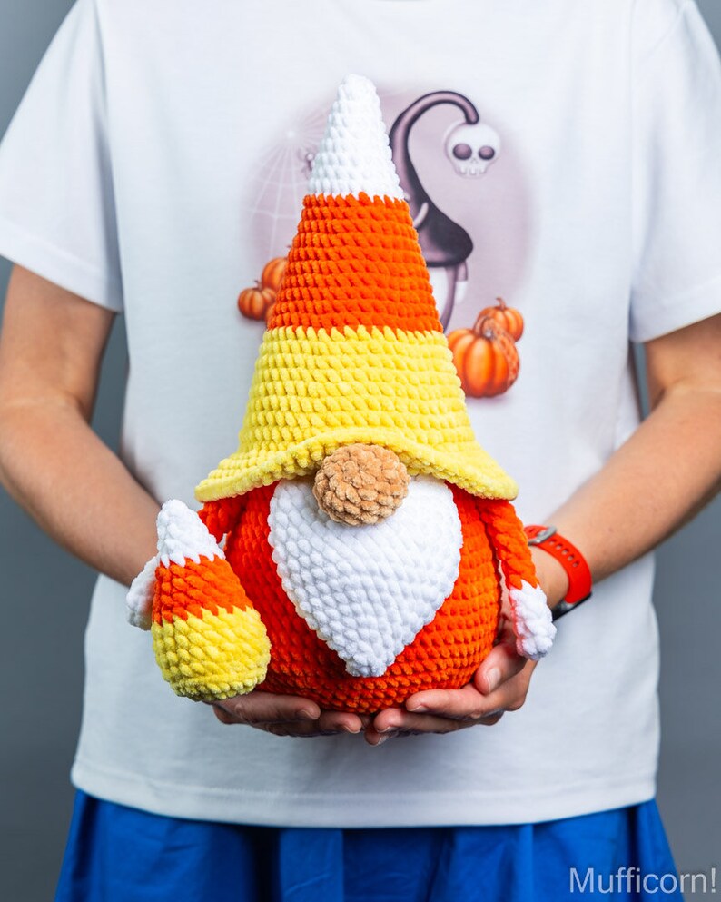 May include: A crocheted candy corn gnome with a white beard and a yellow, orange, and white candy corn hat. The gnome is holding a smaller candy corn gnome in its hands.