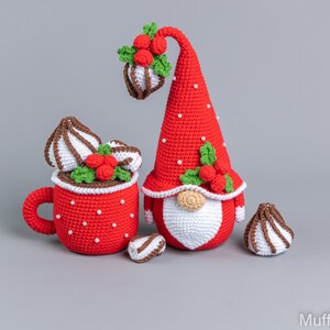 May include: A red crocheted gnome with a white beard and a red hat with holly and berries. The gnome is holding a red crocheted mug with white whipped cream and chocolate sprinkles. There are three additional crocheted whipped cream and chocolate sprinkles on the table.