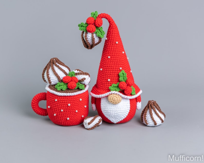 May include: A red crocheted gnome with a white beard and a red hat with holly and berries. The gnome is holding a red crocheted mug with white whipped cream and chocolate sprinkles. There are three additional crocheted whipped cream and chocolate sprinkles on the table.