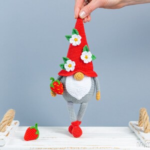 Op de afbeelding: Een gehaakte gnome met een rode muts, witte bloemen en een grijs lichaam. De gnome houdt een rode aardbei in zijn hand. De gnome staat op een wit oppervlak met een touwgreep.