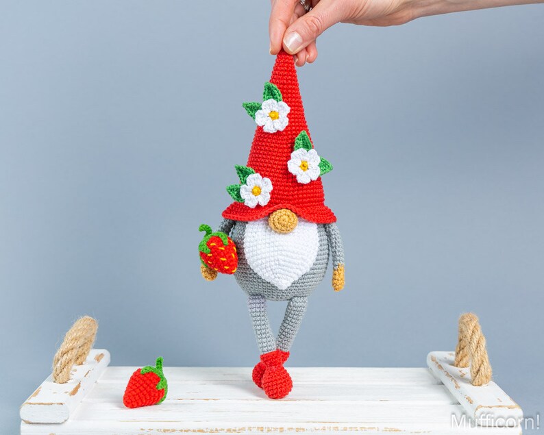 Op de afbeelding: Een gehaakte gnome met een rode muts, witte bloemen en een grijs lichaam. De gnome houdt een rode aardbei in zijn hand. De gnome staat op een wit oppervlak met een touwgreep.