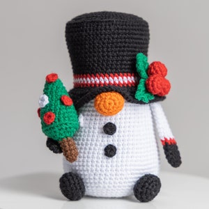 May include: A crocheted snowman holding a small green Christmas tree. The snowman is wearing a black top hat with a red and white band, a white body, black buttons, and a carrot nose. The snowman has black arms and legs.