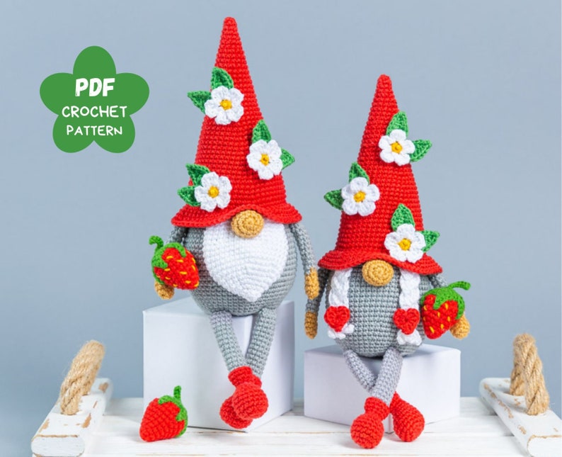 Op de afbeelding: Twee gehaakte gnomen met rode en witte hoeden versierd met bloemen. De gnomen houden aardbeien vast en zitten op een wit oppervlak. PDF Haakpatroon.