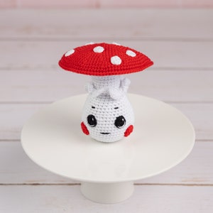 May include: A crocheted mushroom toy with a red cap and white spots. The mushroom has a white body with a cute face and red cheeks.