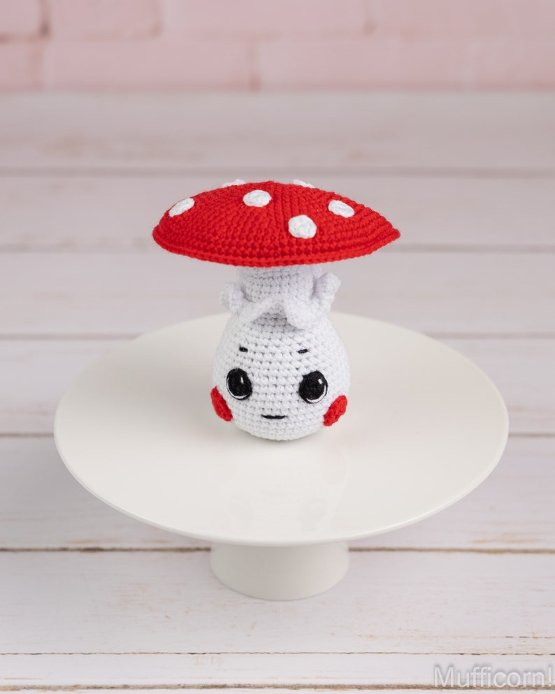 May include: A crocheted mushroom toy with a red cap and white spots. The mushroom has a white body with a cute face and red cheeks.