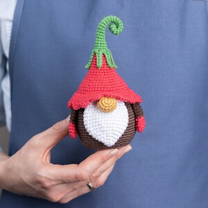 May include: A crocheted gnome with a red hat, brown body, white beard, and green curly hat. The gnome is being held in a person's hand.