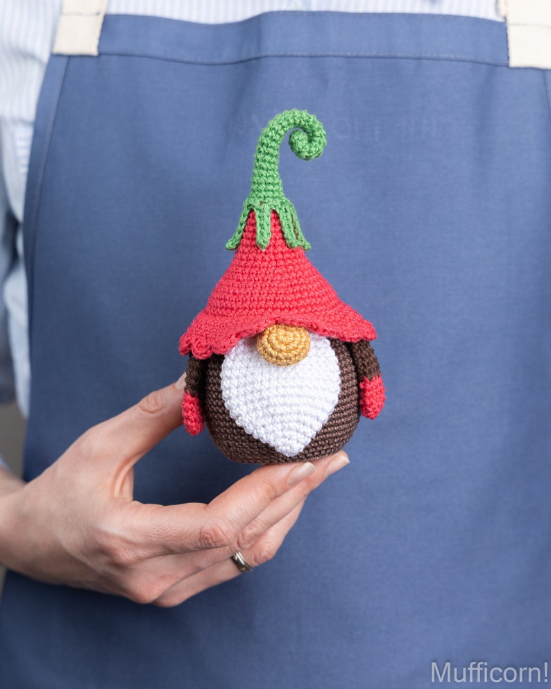 May include: A crocheted gnome with a red hat, brown body, white beard, and green curly hat. The gnome is being held in a person's hand.