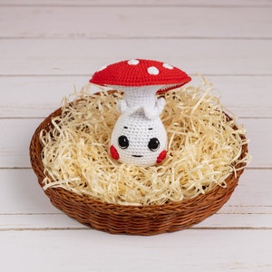 May include: A crocheted mushroom toy with a red and white cap and a white body with a cute face. The mushroom is sitting in a brown wicker basket filled with shredded paper.