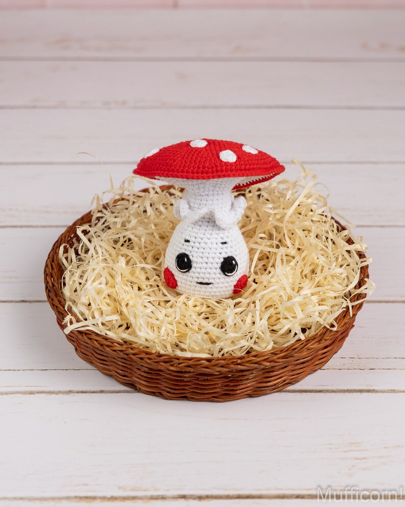 May include: A crocheted mushroom toy with a red and white cap and a white body with a cute face. The mushroom is sitting in a brown wicker basket filled with shredded paper.