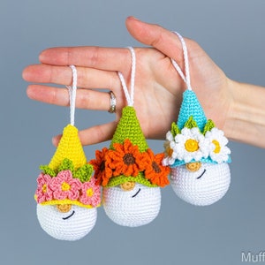 May include: Three crocheted gnome ornaments with different colored hats and flower accents. The gnomes are white with black eyes and a smiling mouth. The hats are yellow, green, and blue with flowers in shades of pink, orange, and white.