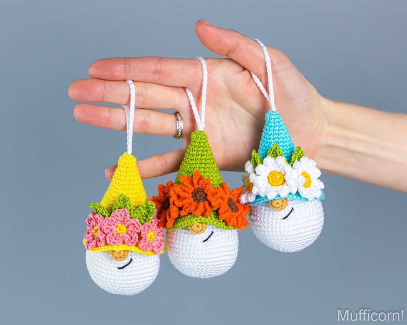 May include: Three crocheted gnome ornaments with different colored hats and flower accents. The gnomes are white with black eyes and a smiling mouth. The hats are yellow, green, and blue with flowers in shades of pink, orange, and white.