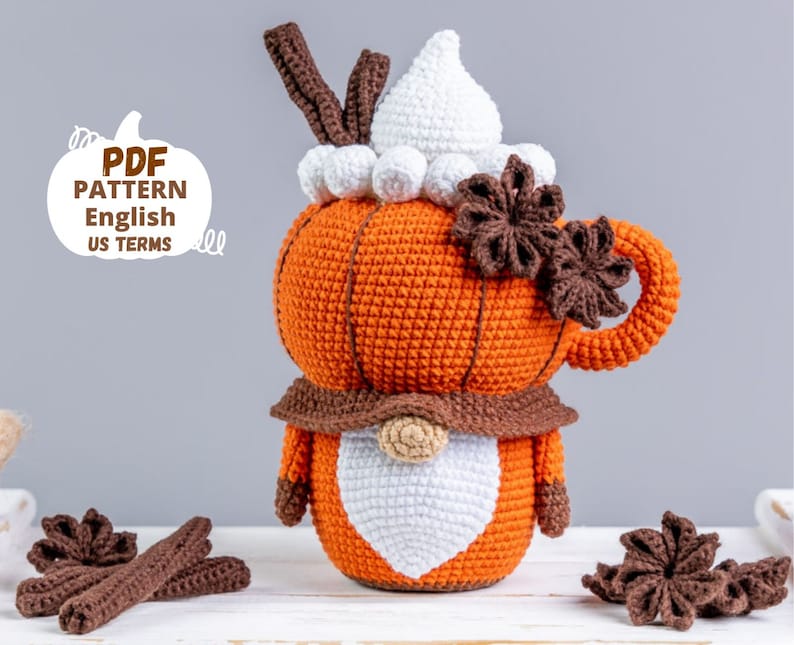 Puede incluir: Un gnomo con forma de calabaza tejido a ganchillo, con barba y sombrero blancos, cuerpo naranja y detalles marrones. El gnomo sostiene una taza tejida con una cobertura de crema batida blanca y an&iacute;s estrellado marr&oacute;n. El texto "PDF PATTERN English US TERMS" est&aacute; a la izquierda.