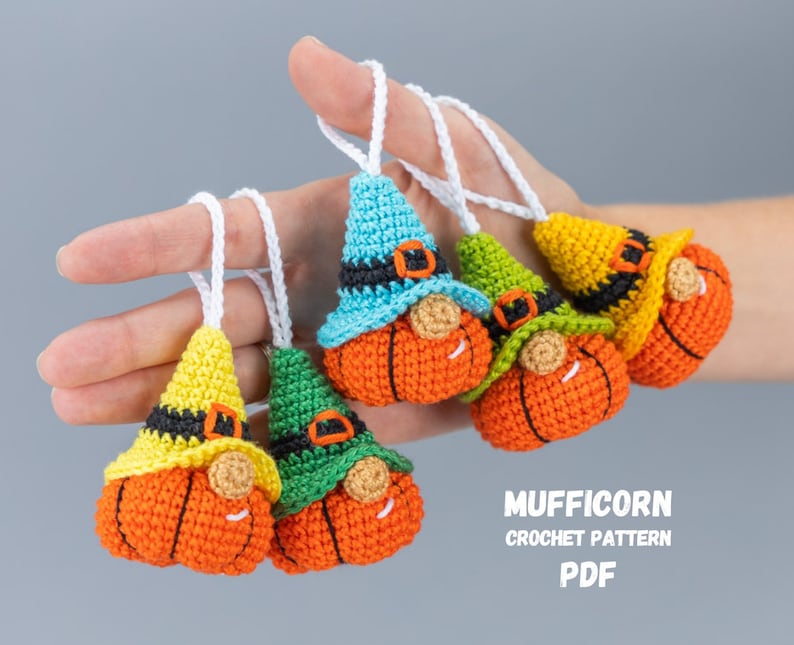 May include: Five crocheted pumpkin gnome ornaments with different colored hats. The gnomes are wearing black belts and have orange pumpkin bodies. The hats are blue, green, yellow, and two are yellow. The gnomes are hanging from white strings. The text "MUFFICORN CROCHET PATTERN PDF" is at the bottom of the image.