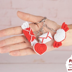 May include: A set of crocheted charms featuring a heart, a candy, and two envelopes. The charms are attached to a silver key ring. The charms are red and white.