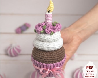 May include: A crocheted cupcake toy with a pink and white frosting, brown cupcake base, and a pink and white striped candle with a yellow flame. The cupcake is decorated with pink roses and green leaves.