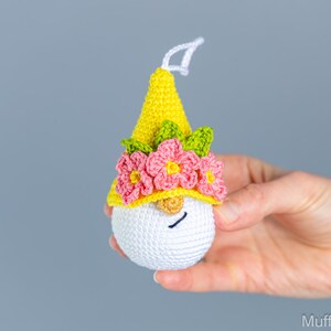 May include: A crocheted gnome ornament with a yellow hat, pink flowers, and green leaves. The gnome has a white face with a black smile.